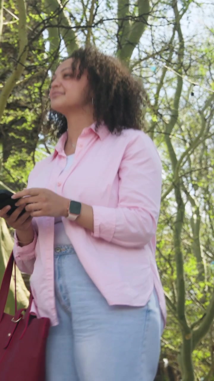 Video verticale: Donna con camicia rosa che tocca lo smartphone, controllando i messaggi su un viale del parco, con una borsa rossa.