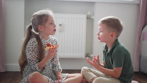 smiling girl eating grilled chicken pizza slice while sitting on floor talking with brother in bright sunlit cozy room, laughing together as girl gently removes something from her eye