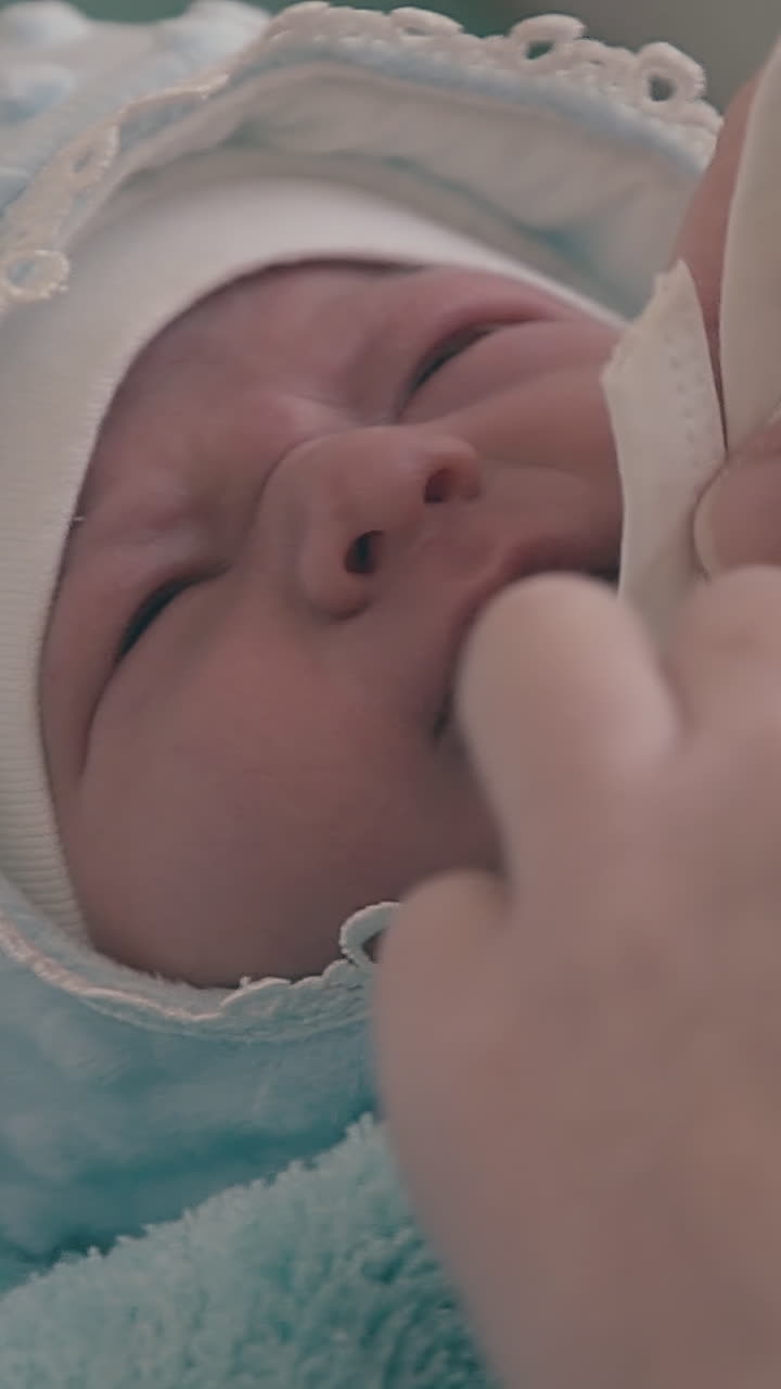 young mommy ties ribbons of warm bonnet on crying newborn baby head by hands in light room extreme close view