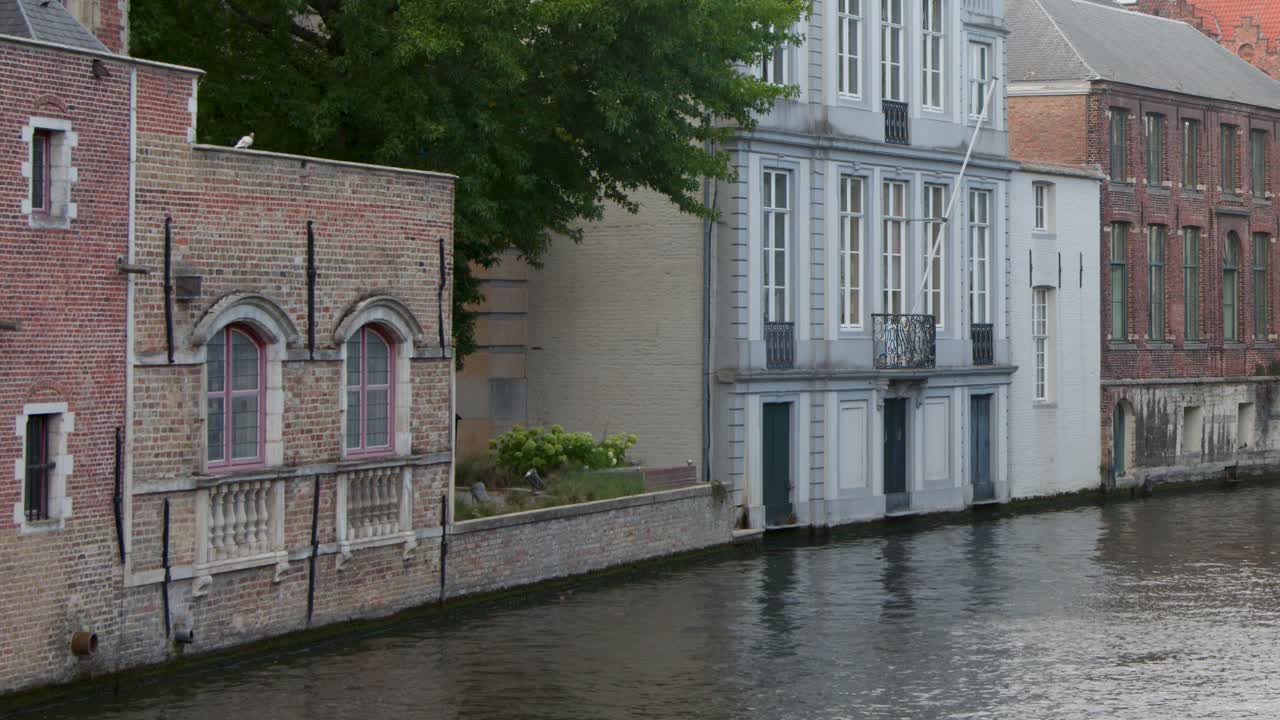 Smooth camera pan along tranquil Bruges canal, historic riverside buildings, autumn foliage, overcast daylight