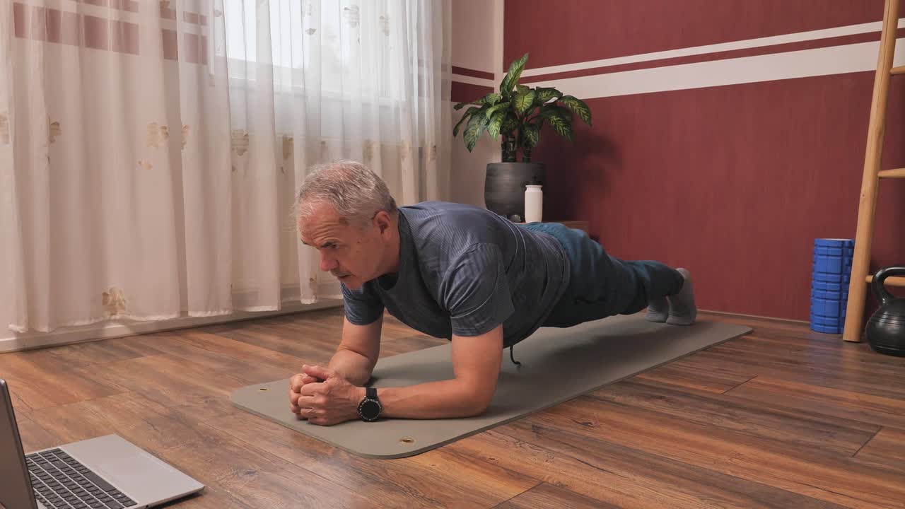 Man doing plank on yoga mat at home, following online workout in bright fitness space