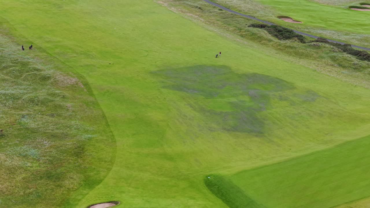 Drone glides above lush links golf course, golfers visible, overcast daylight, wide aerial perspective
