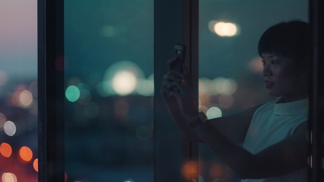 아시아의 사업가 여성이 스마트폰을 사용하여 밤에 도시의 사진을 찍고 아름다운 전망을 즐기며 사무실에서 늦게 일하고 있습니다.