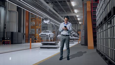 cuerpo lleno de un trabajador profesional masculino asiático de pie con su teléfono inteligente en la fábrica de automóviles ev, instalación industrial de alta tecnología, trabajando continuamente
