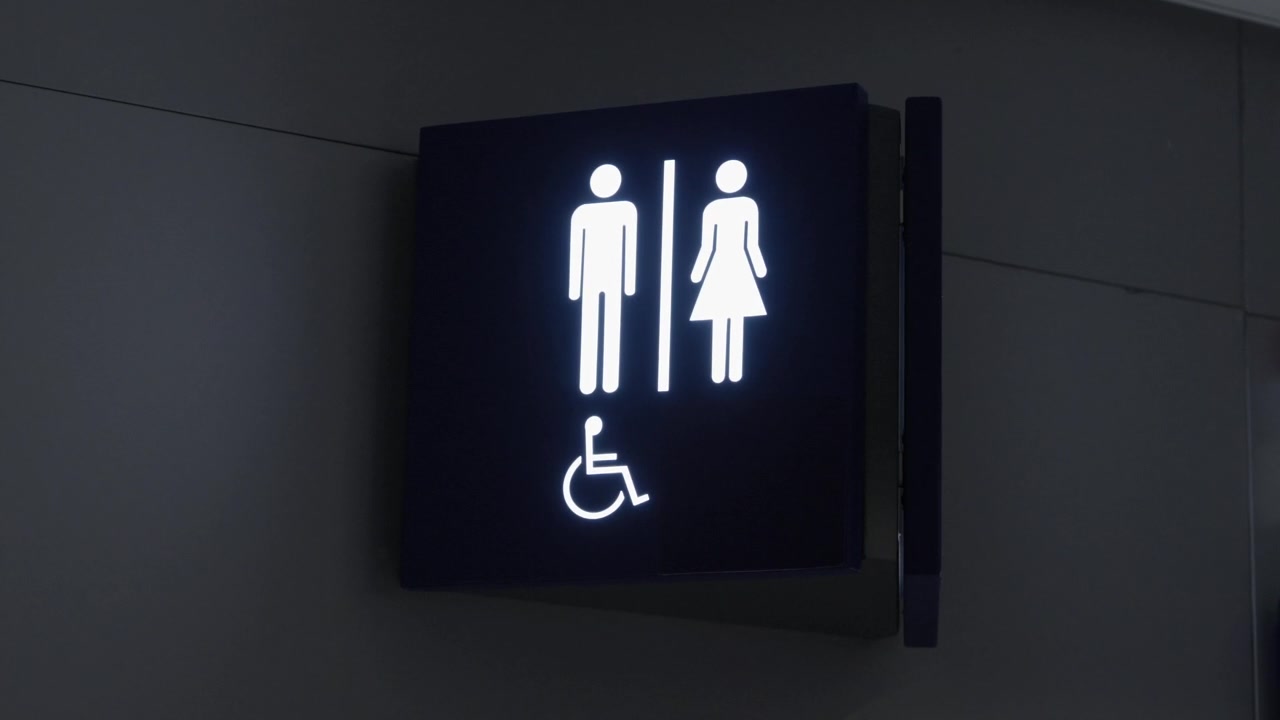 Illuminated public toilet sign with male female and wheelchair accessible symbols mounted on a dark wall