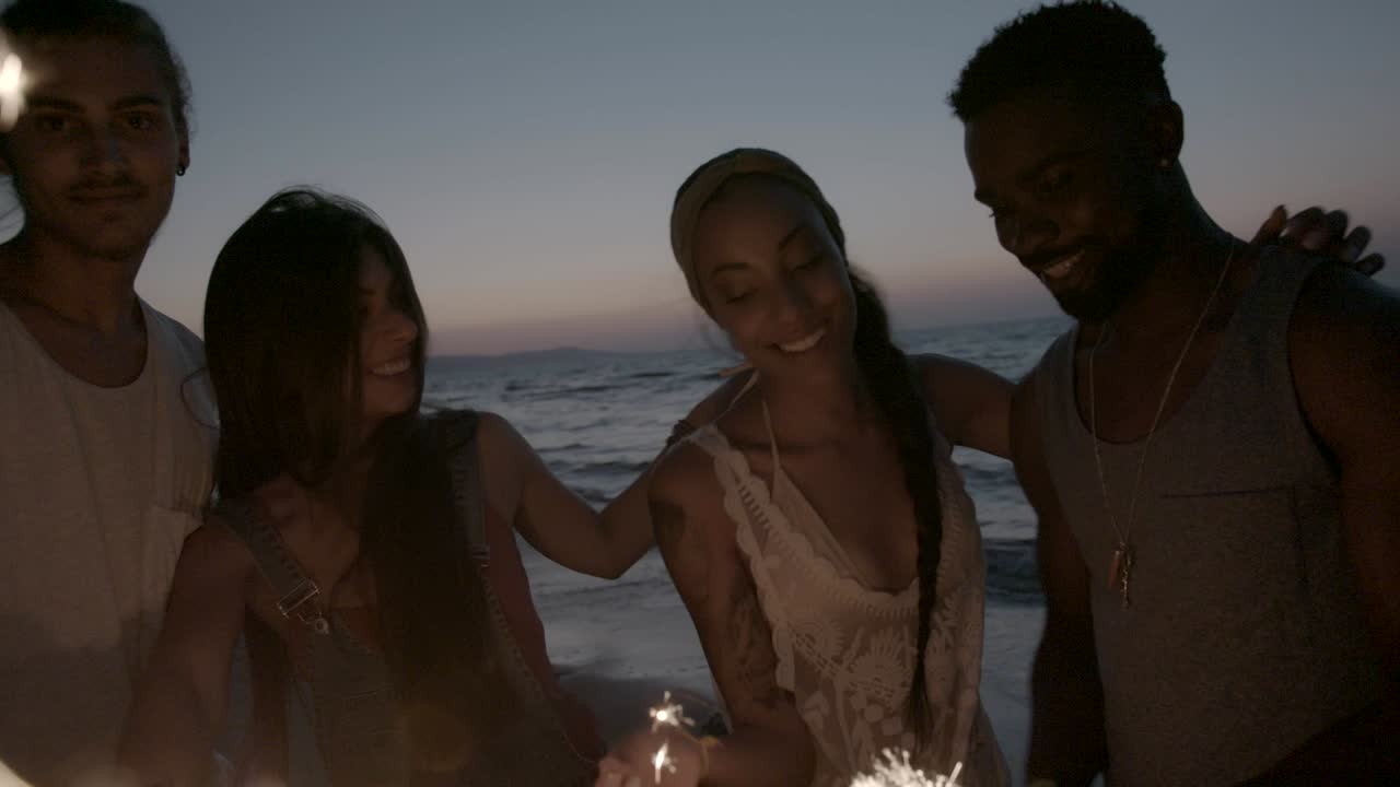 다민족 커플들이 밤에 해변에서 반이는 불꽃으로 축하하고 있습니다.