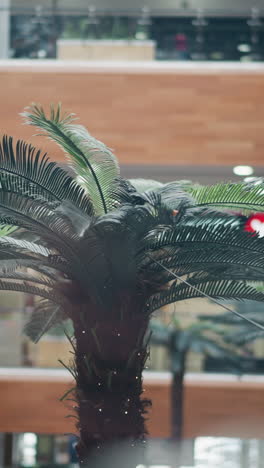 close-up of artificial palm tree inside modern shopping mall, surrounded by blurred retail storefronts and warm interior tones, showcasing vibrant greenery and architectural aesthetics