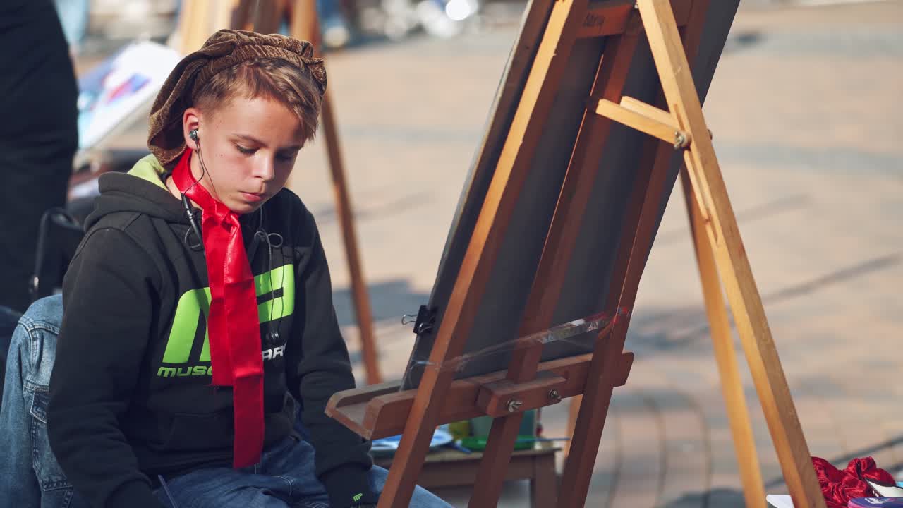 Serious boy is painting on easel in the city. Portrait of a kid artist practices his skills in drawing with brush on the busy city background. City fair.