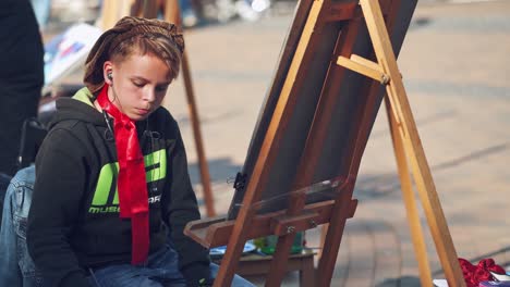 serious boy is painting on easel in the city. portrait of a kid artist practices his skills in drawing with brush on the busy city background. city fair.