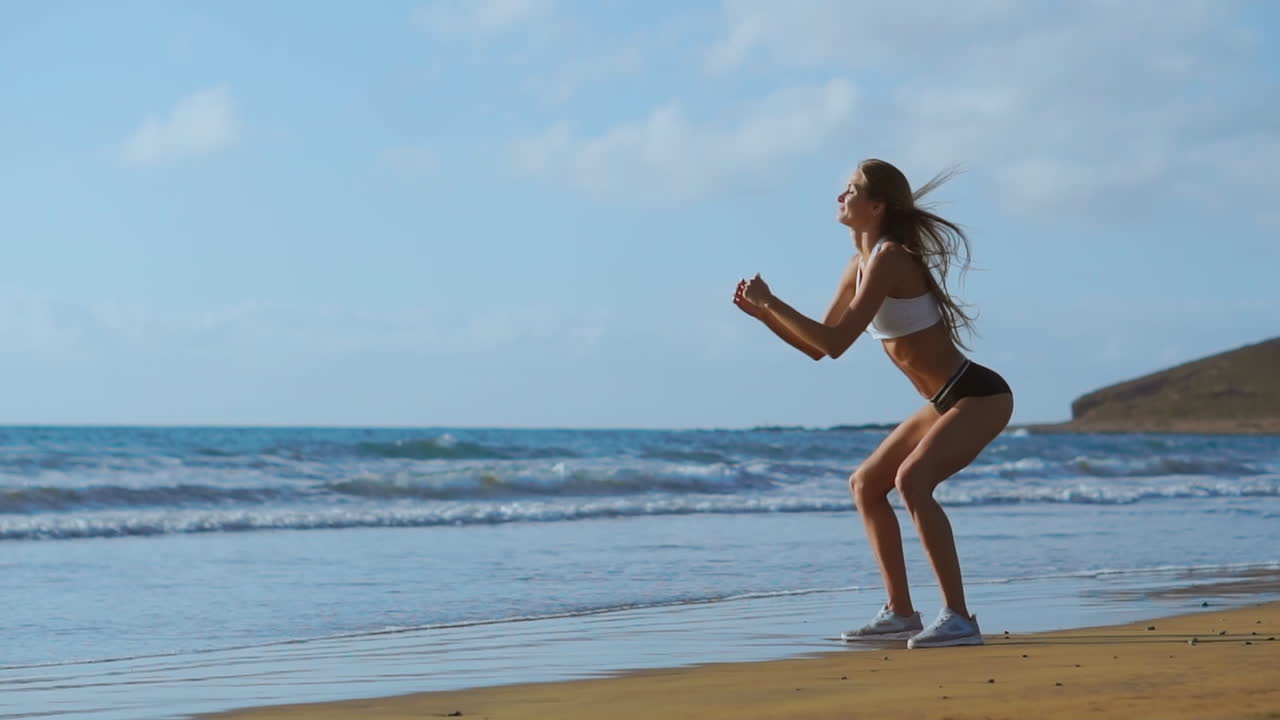 Giovane donna in forma che allena core e glutei con un allenamento a corpo libero, facendo squat in spiaggia. Ragazza sportiva che piega le gambe come parte di una vita attiva e in forma. Ripresa Steadicam.