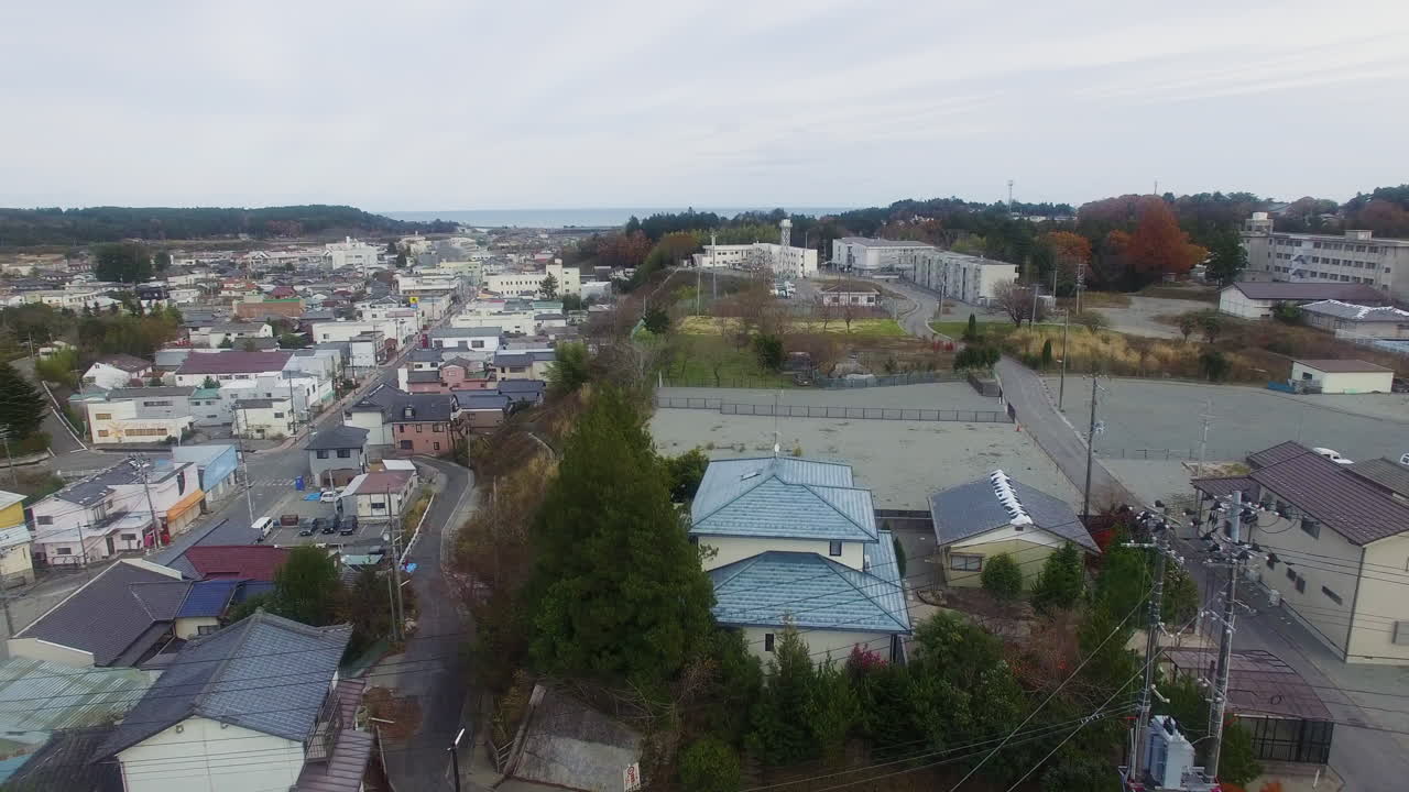 Drone footage captures an aerial view of a suburban neighborhood in Fukushima Prefecture, Japan. The scene depicts quiet streets and houses under a somber sky, highlighting the atmospheric stillness o