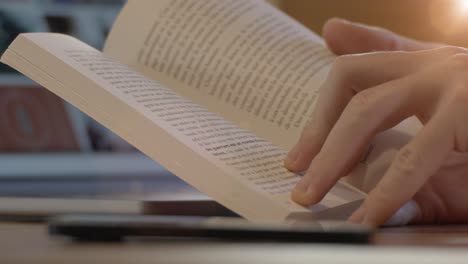 primer plano: hombre absorto en la lectura de un libro de bolsillo, volteando páginas, smartphone borroso en primer plano