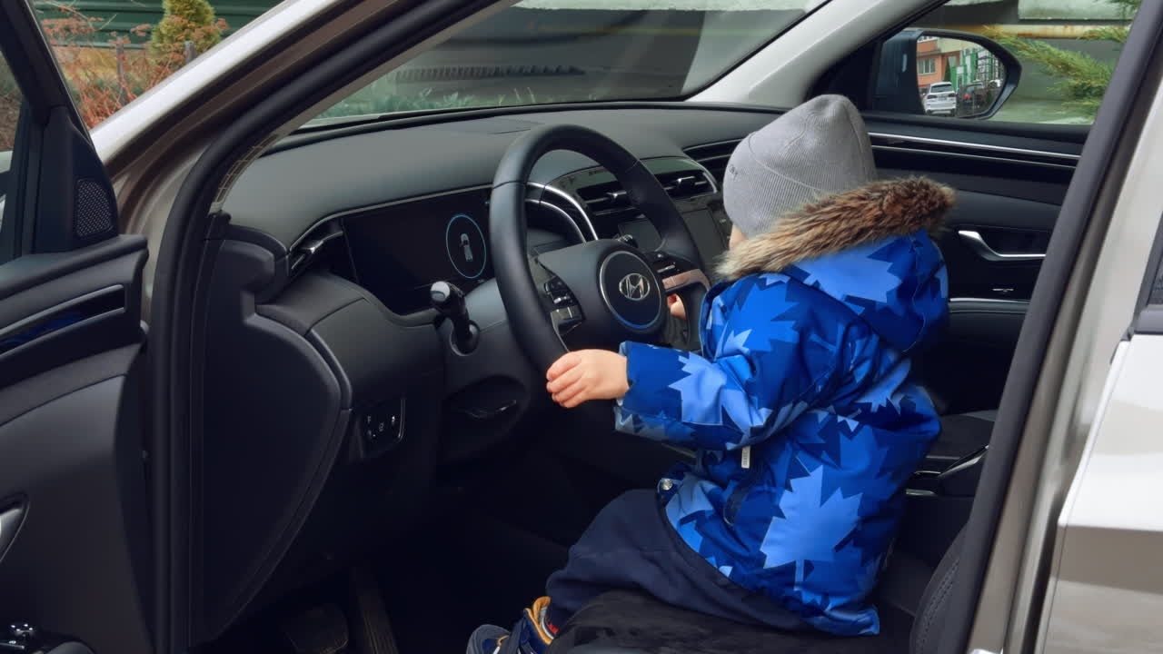 Male kid in blue jacket sits on the driver's seat of the car. Side view of a baby boy holding by the steering wheel.