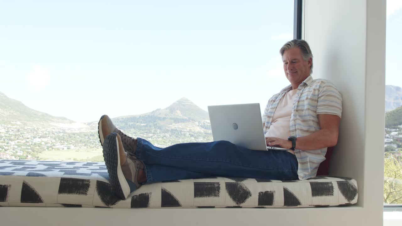Mature man placing laptop on lap, reclining and typing on window seat at home, working remotely