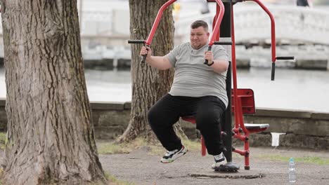 a young overweight man is working out doing exercises on outdoor exercise machine in a green area a young overweight man is working out doing exercises on an outdoor exercise machine in a green area a fat man struggles with excess weight exercises for hands