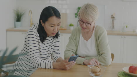 woman helping elderly woman with smartphone in kitchen