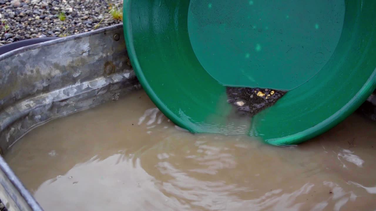Gold panning black silt, sand, clay, and gravel alluvial deposits in green trap pan, wetting, soaking, agitating and washing in brown water in tin bucket, looking at nuggets, static close up portrait