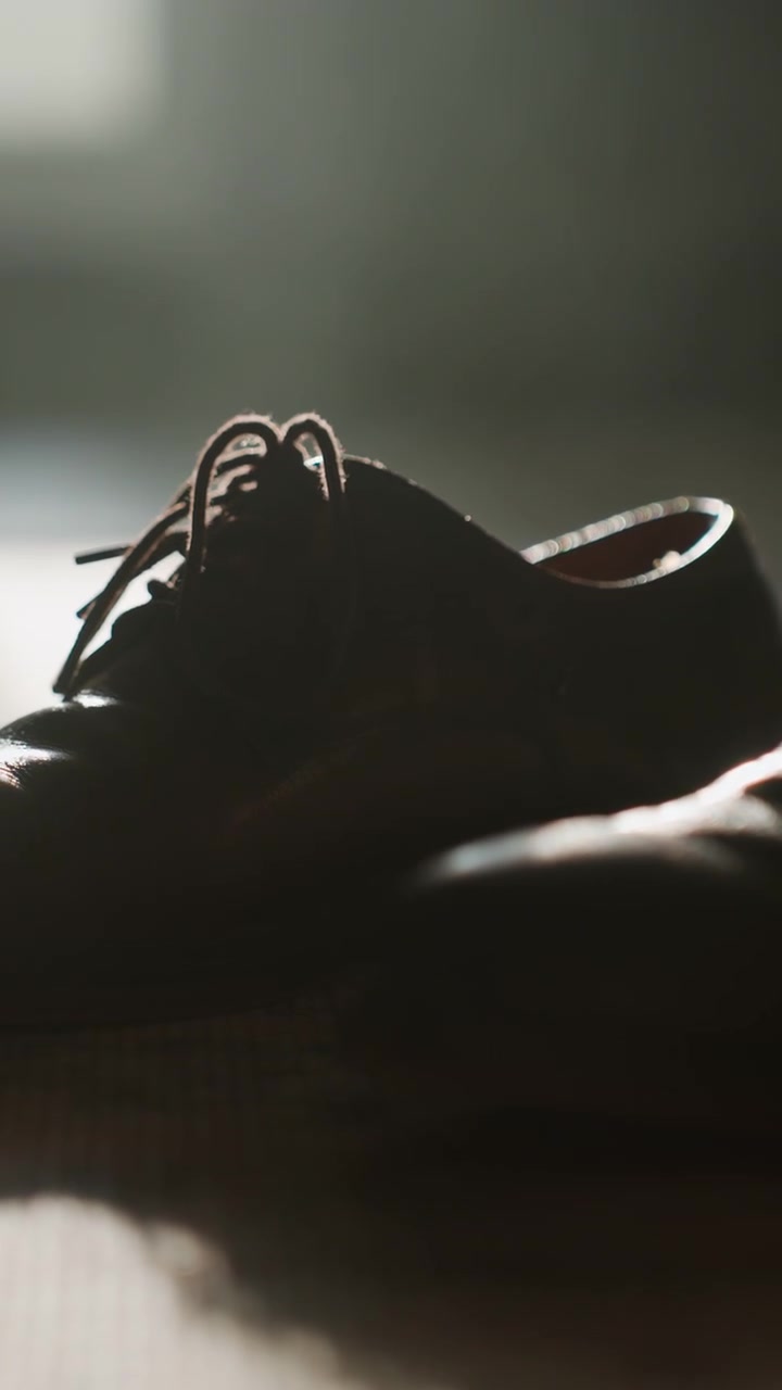 Scarpe Oxford in pelle illuminate dal sole sul pavimento, brogue lucide che catturano la calda luce della finestra. Lunghe ombre morbide su un tappeto testurizzato, primo piano di lacci e puntale. Profondità di campo cinematografica, un tranquillo rituale di vestizione.