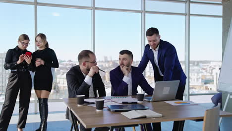 men in office talking about work sitting at the desk with papers and laptop. business ladies standing at the backdrop. panoramic windows with city view at the backdrop.