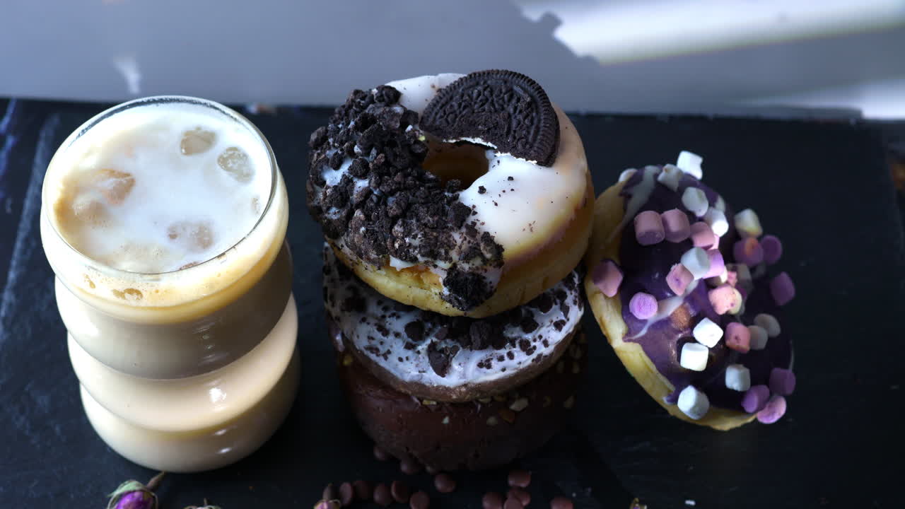 A hand with pink nails lifts a white-glazed donut topped with chocolate cookie crumbles and a sandwich cookie from a colorful stack. Iced coffee, a marshmallow donut complete the cozy slate tabletop