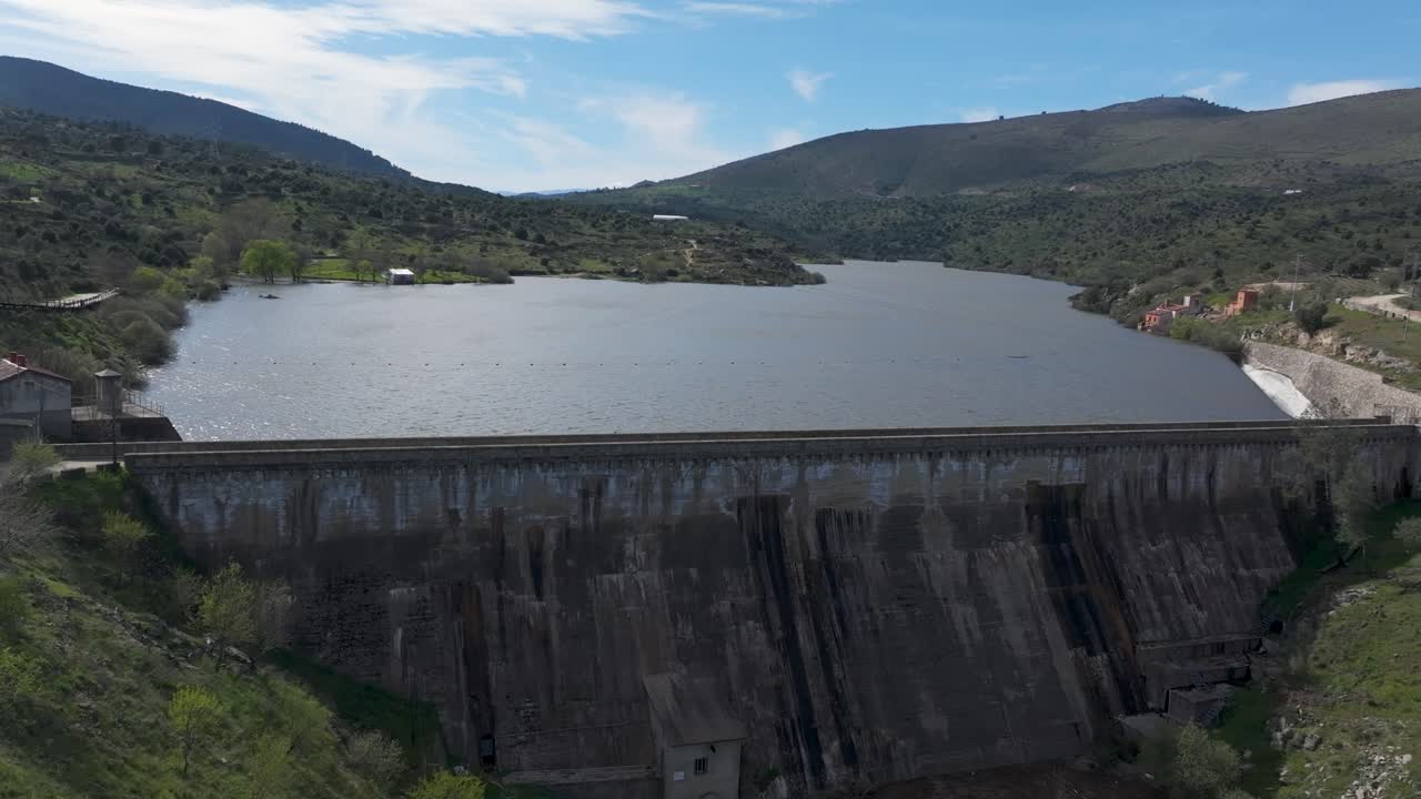 Ascending drone footage reveals the towering dam wall, with a lateral spillway releasing water. The reservoir is completely full, and the climb unveils the surrounding scenic environment.