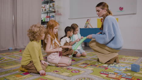 four children sitting on a carpet while their teacher helps them inflate long balloons in a montessori school