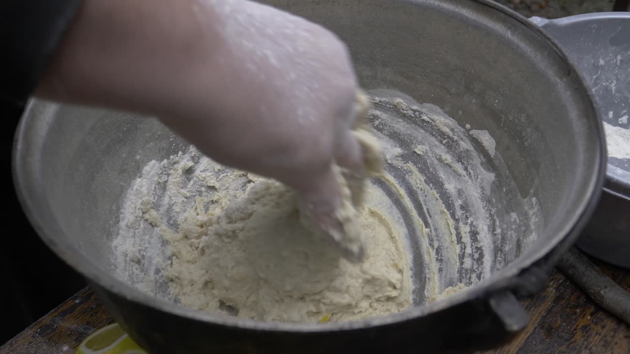 Kneaded dough in a black cauldron. The dough is soft and covered with flour, while it is kneaded with one hand by a chef in nature. The cauldron is on a chopper above a table.