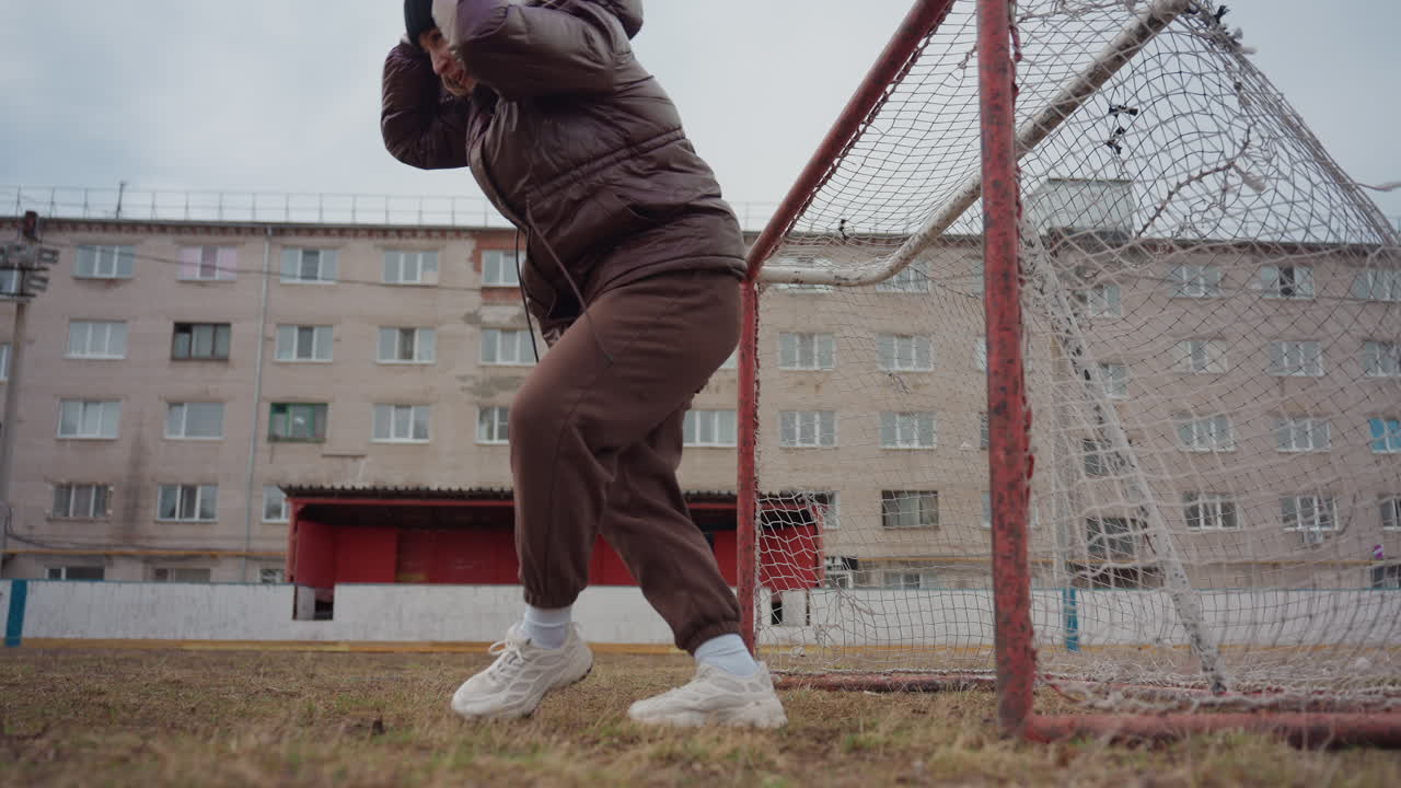 Portero preparándose en la portería con una mujer mayor de pelo blanco revisando la red y su posición; el ángulo bajo de la cámara captura la postura y los zapatos; bloques de apartamentos detrás, clima nublado, calentamiento tranquilo y concentrado