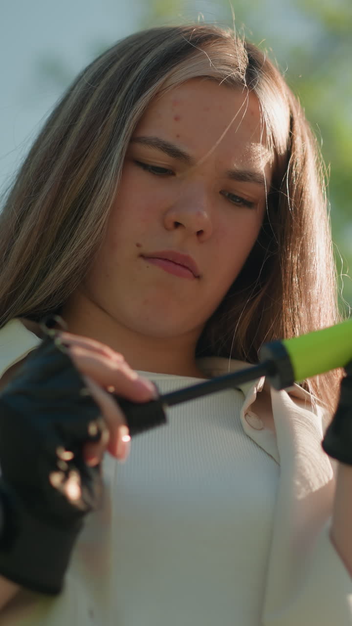 mujer enfocada con cabello rubio se encuentra al aire libre, luchando para ajustar la boquilla de la bomba de aire verde en sus manos con guantes, en un fondo natural de árboles verdes exuberantes