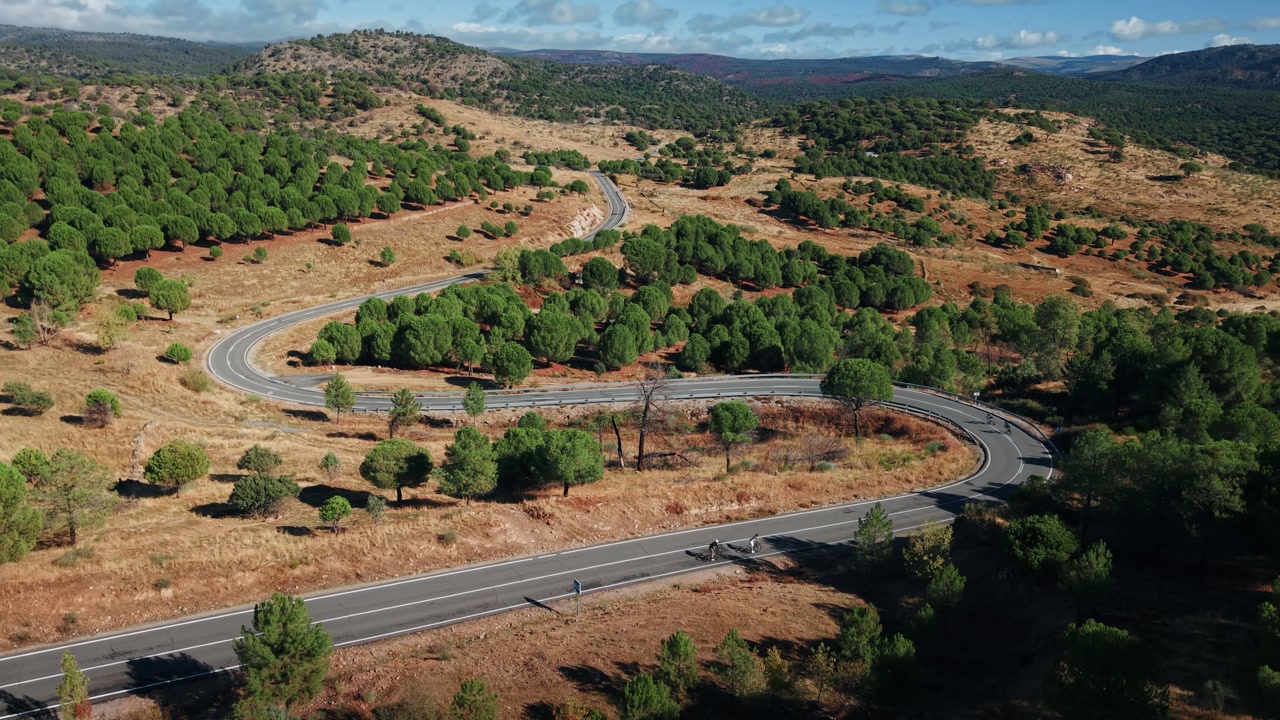 Drone pulling back over winding road in Madrid countryside with cyclists riding through dry landscape and hills.