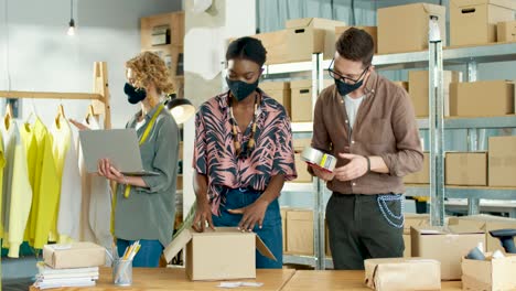 caucasian young man and african american woman working in clothing shop packing parcels while caucasian woman typing on laptop