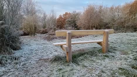 Leere-Hölzerne-Parkbank-Auf-Frostigem,-Eisbeschichtetem-Grashang-Mit-Blick-Auf-Herbstliches-Waldlaub