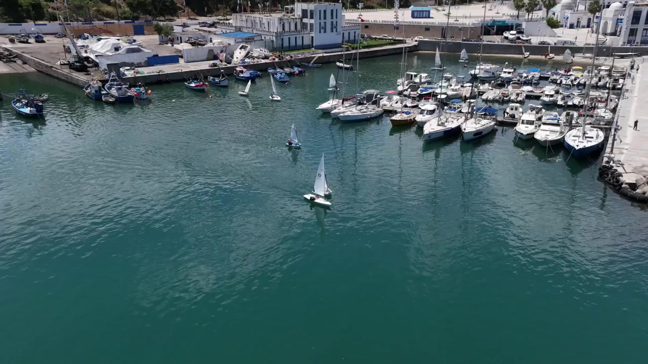 Vista aerea di piccole barche a vela che si muovono all'interno di un porto turistico mediterraneo nel Marocco settentrionale, circondate da yacht ormeggiati, strutture portuali e acque costiere calme