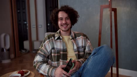 Portrait-of-a-happy-brunette-guy-with-curly-hair-in-a-checkered-shirt-who-holds-a-smartphone-in-his-hands-and-sits-on-a-comfortable-chair-in-a-modern-cozy-apartment