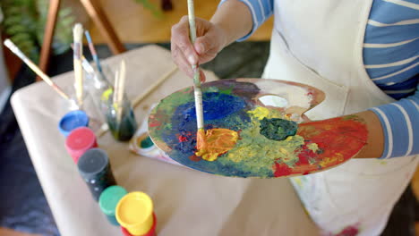 Senior-biracial-woman-wearing-apron-and-mixing-colours-on-palette-at-home,-slow-motion