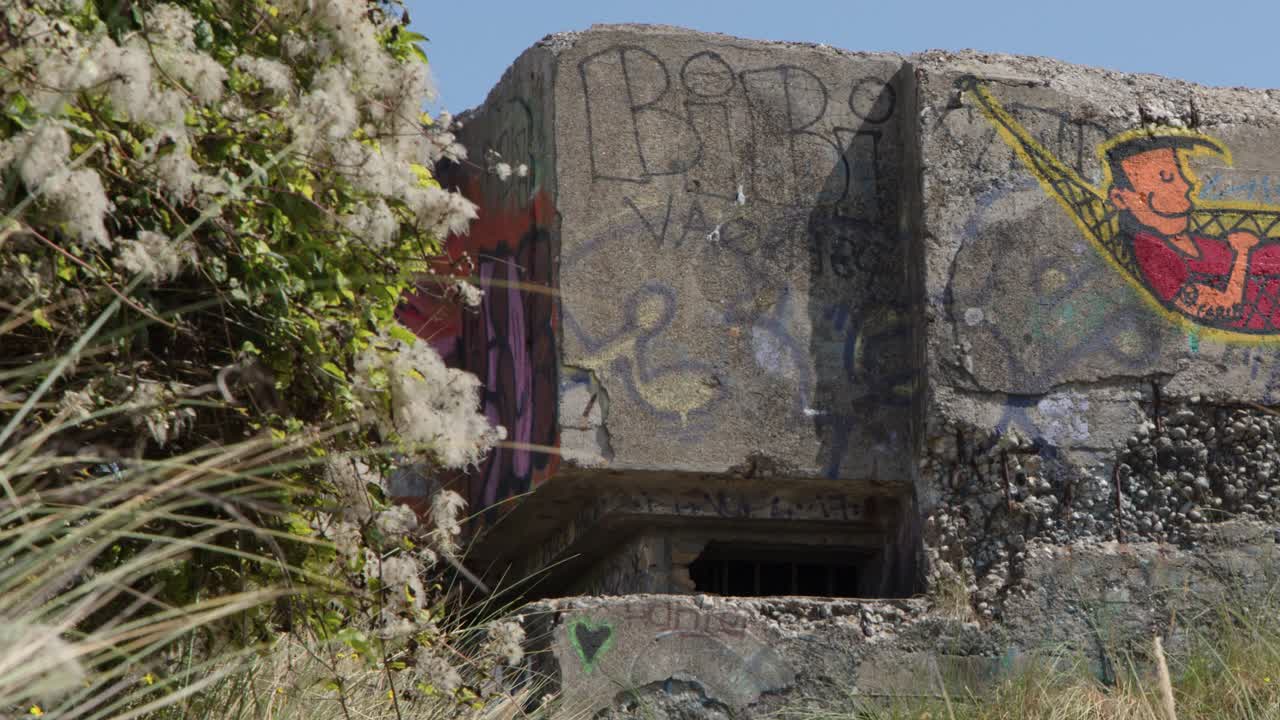 Daylight pan reveals abandoned concrete bunker with colorful street art amid grassy seaside dunes