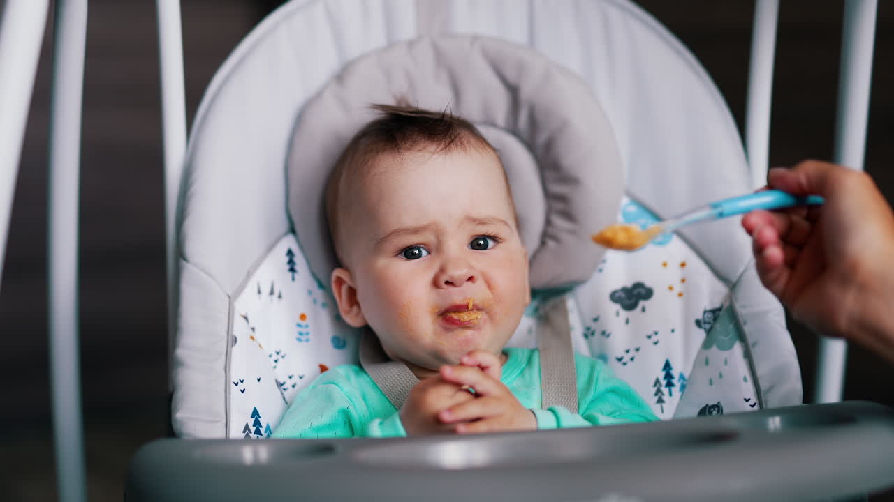 Baby feeding from spoon. Mother's hand gives the orange stuff to her son and he makes faces after.