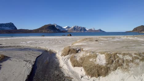 Toma-Aérea-Con-Plataforma-Rodante-Sobre-Las-Dunas-De-Arena-En-La-Famosa-Playa-De-Haukland-En-Lofoten,-Noruega,-A-Finales-Del-Invierno-Con-Pequeños-Parches-De-Nieve-E-Impresionantes-Vistas-A-Las-Montañas-Sobre-El-Agua.