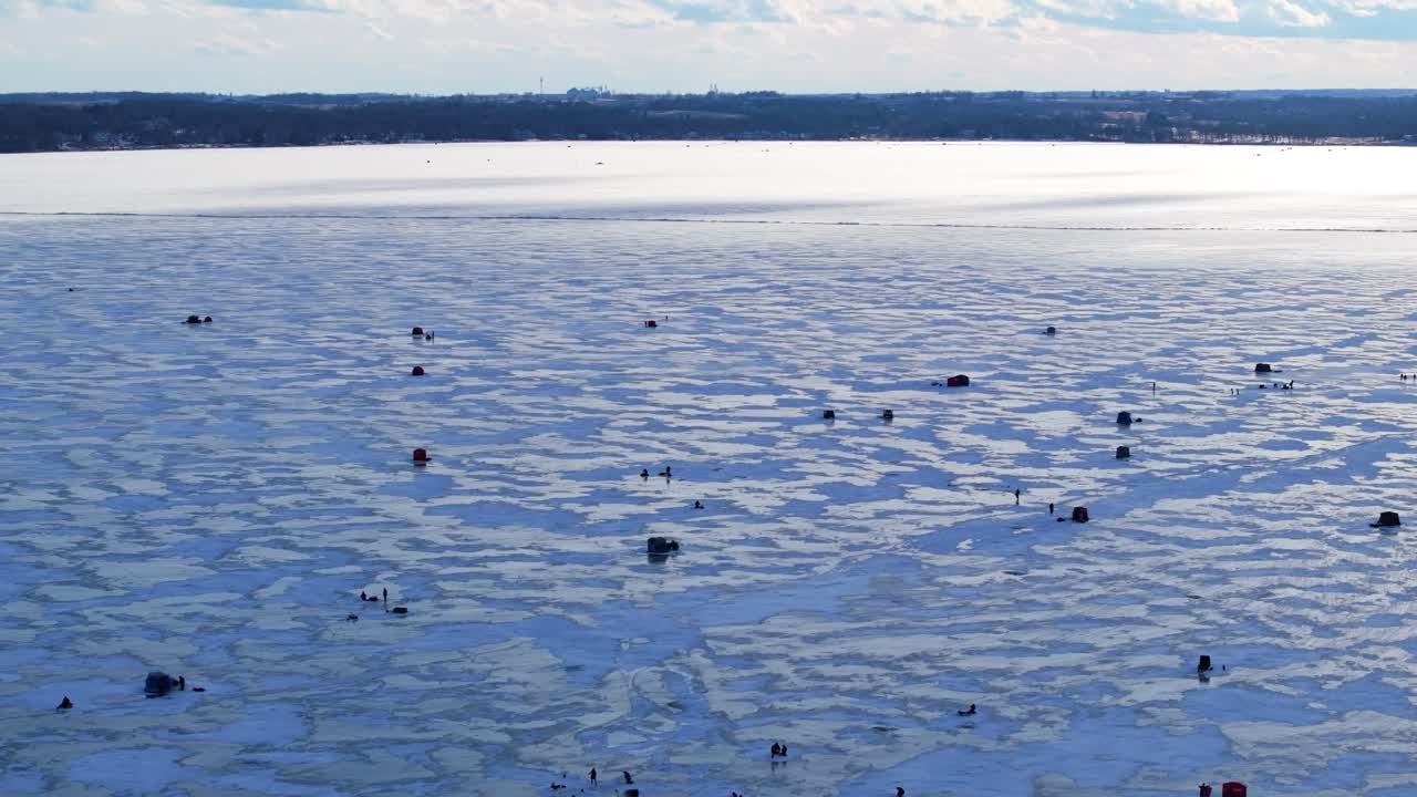 Frozen lake surface with ice fishing tents and blowing snow across textured cracked surface, drone at golden hour
