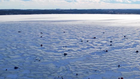 frozen lake surface with ice fishing tents and blowing snow across textured cracked surface, drone at golden hour