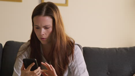 Anxious-Woman-Sitting-On-Sofa-At-Home-Looking-At-Mobile-Phone-Concerned-About-Social-Media-Or-Bad-News