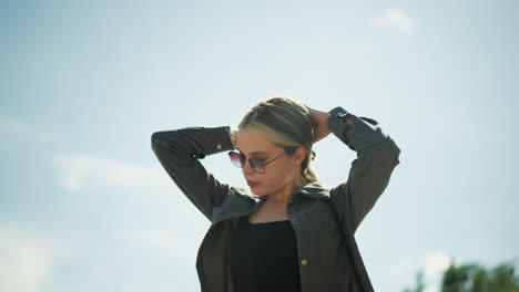 woman wearing black top under grey jacket and sunglasses gently touches her hair as the wind blows, under the bright, sunny sky, with her hair fluttering in the breeze