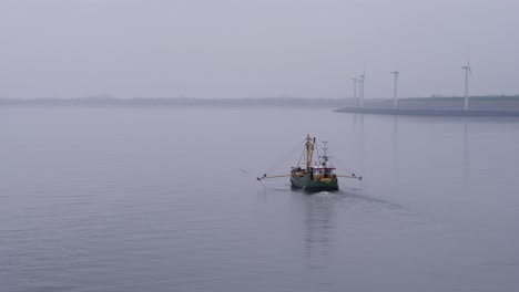 green fishing boat with outstretched yellow fishermen's arms sails over the calm north sea with the zeeland dikes with modern windmills in the background on a misty day