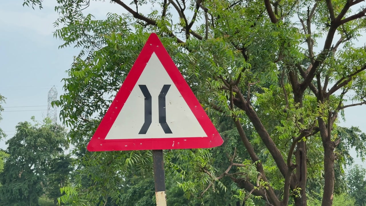 A triangular road sign indicating road narrowing ahead, captured in a static shot with trees in the background.