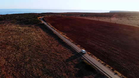 Aerial-track-shot-of-lonely-van-driving-on-portguese-countryside-road-with-beautiful-ocean-in-background