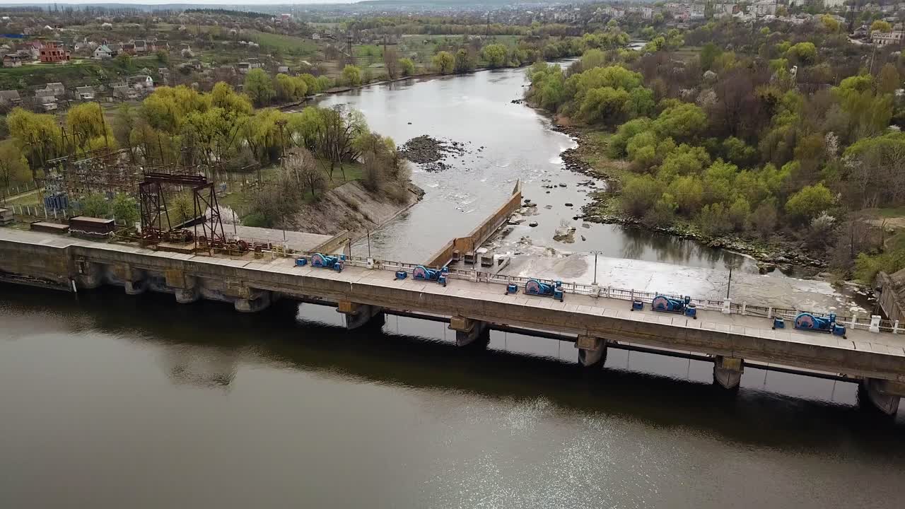 A huge construction with a bridge stands on the river outside the city for water purification on the background of wonderful landscape of green trees and with buildings. Close-up. Aerial view.