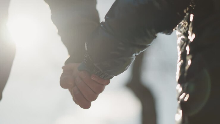 A close-up of an adult and child holding hands, with sunlight reflecting off their hands, the child is wearing a black jacket