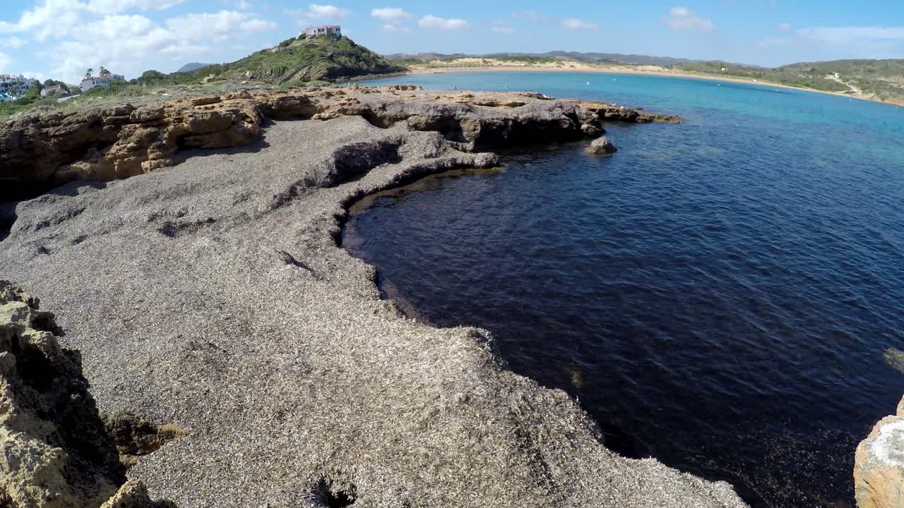Rocky shoreline covered in seaweed at Cala Tirant, Menorca, with clear blue waters