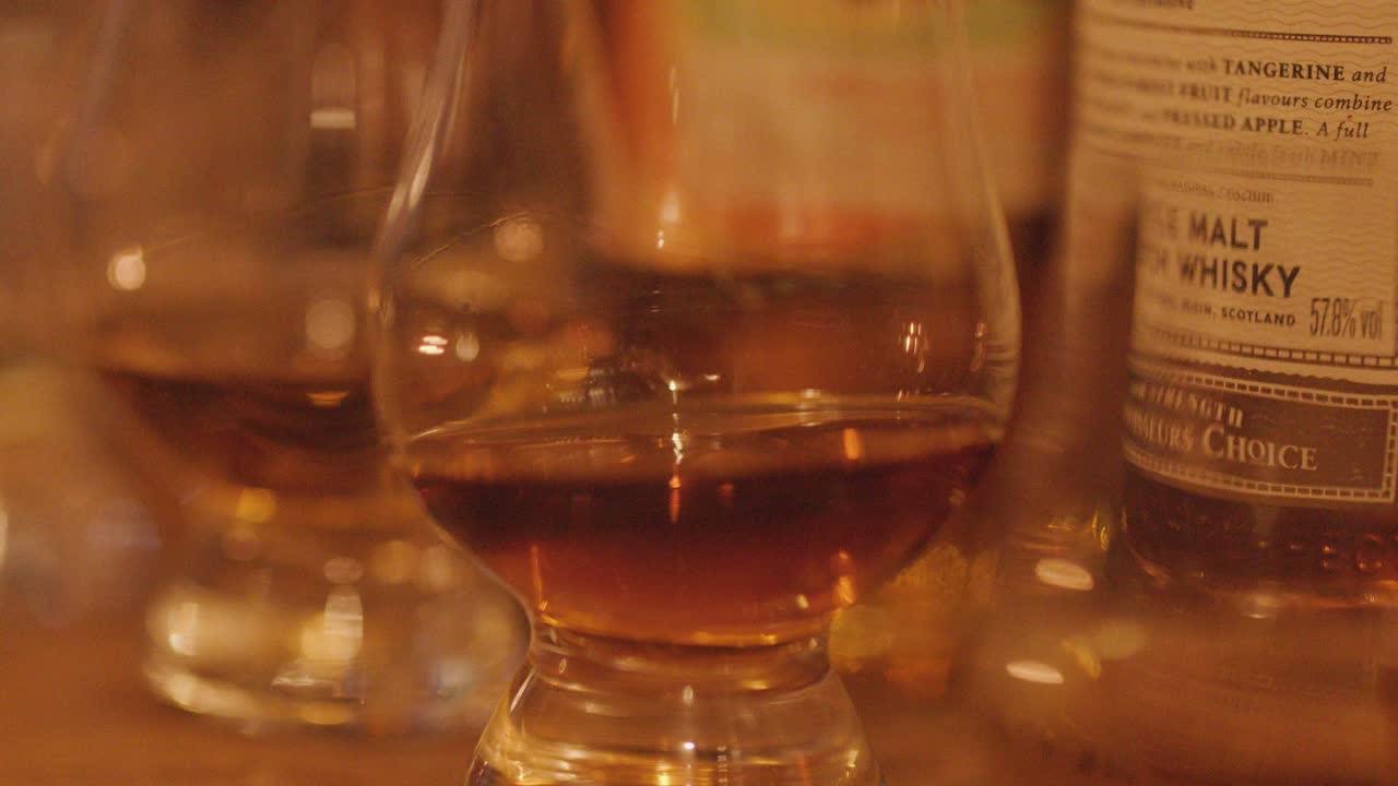A hand lifts a whisky glass from a wooden bar, surrounded by bottles and glasses, under warm ambient lighting with a shallow depth of field