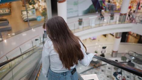 rear view of young adult flaunting her hair on descending escalator with hand on rail in busy mall, capturing movement and casual elegance in a public shopping space